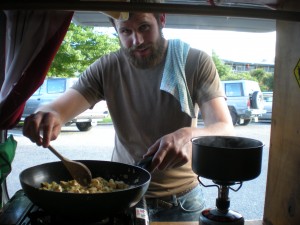 Tall man cooking food in the back of a hippie van - creating a good cooking set up is a big part of figuring out how to live in a van
