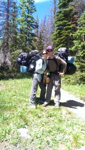 couple standing by a trail in the trees wearing hiking packs with an excessive amount of gear attached