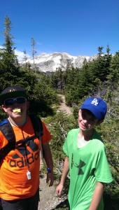 two boys in brightly colored tshirts standing on a nature trail with snowy and rocky mountains in background