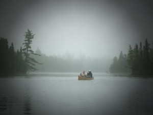 couple paddling in the BWCA canoe on lake surrounded by trees with dark grey skies and lots of mist/fog